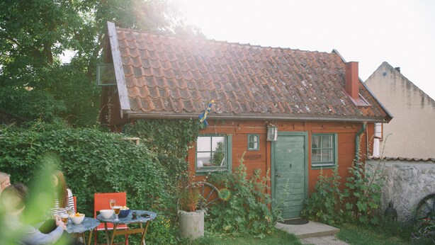 Ett litet hus med ett tak täckt av tegelpannor, en grön dörr och ett fönster. Utanför finns ett bord med stolar, och en krukväxt står framför huset.