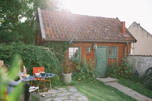 Una casa pequeña con techo de tejas, una puerta verde y una ventana. Hay una mesa con sillas afuera, y una planta en maceta frente a la casa.
