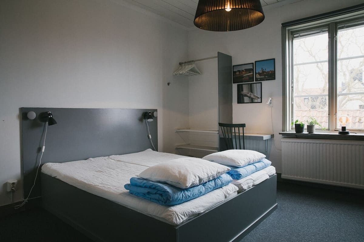 Een slaapkamer met een bed, een raam met planten, een radiator en een hanglamp.