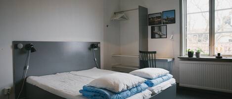 A bedroom with a bed, a window with plants, a radiator, and a hanging light fixture.