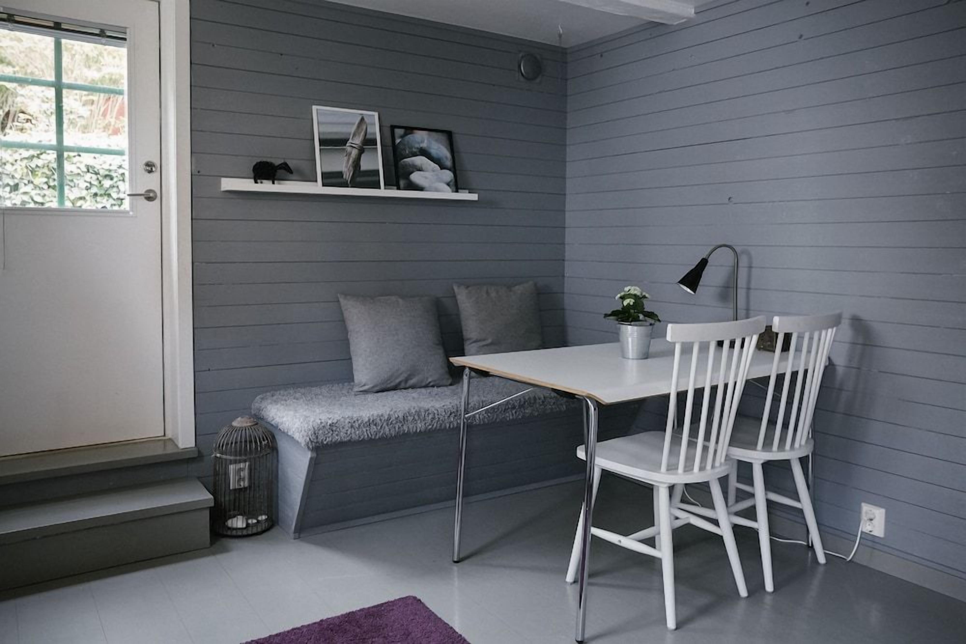 Una habitación pequeña y moderna con una pared gris, una puerta blanca, un banco gris con cojines, una mesa blanca con una lámpara y una silla blanca.