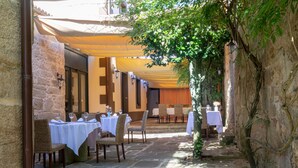 An outdoor dining area with stone flooring, tables with white tablecloths, wicker chairs, and a yellow canopy providing shade. - Hotel Restaurante Asador de Roxos Casa de Albardonedo (Santiago de Compostela)