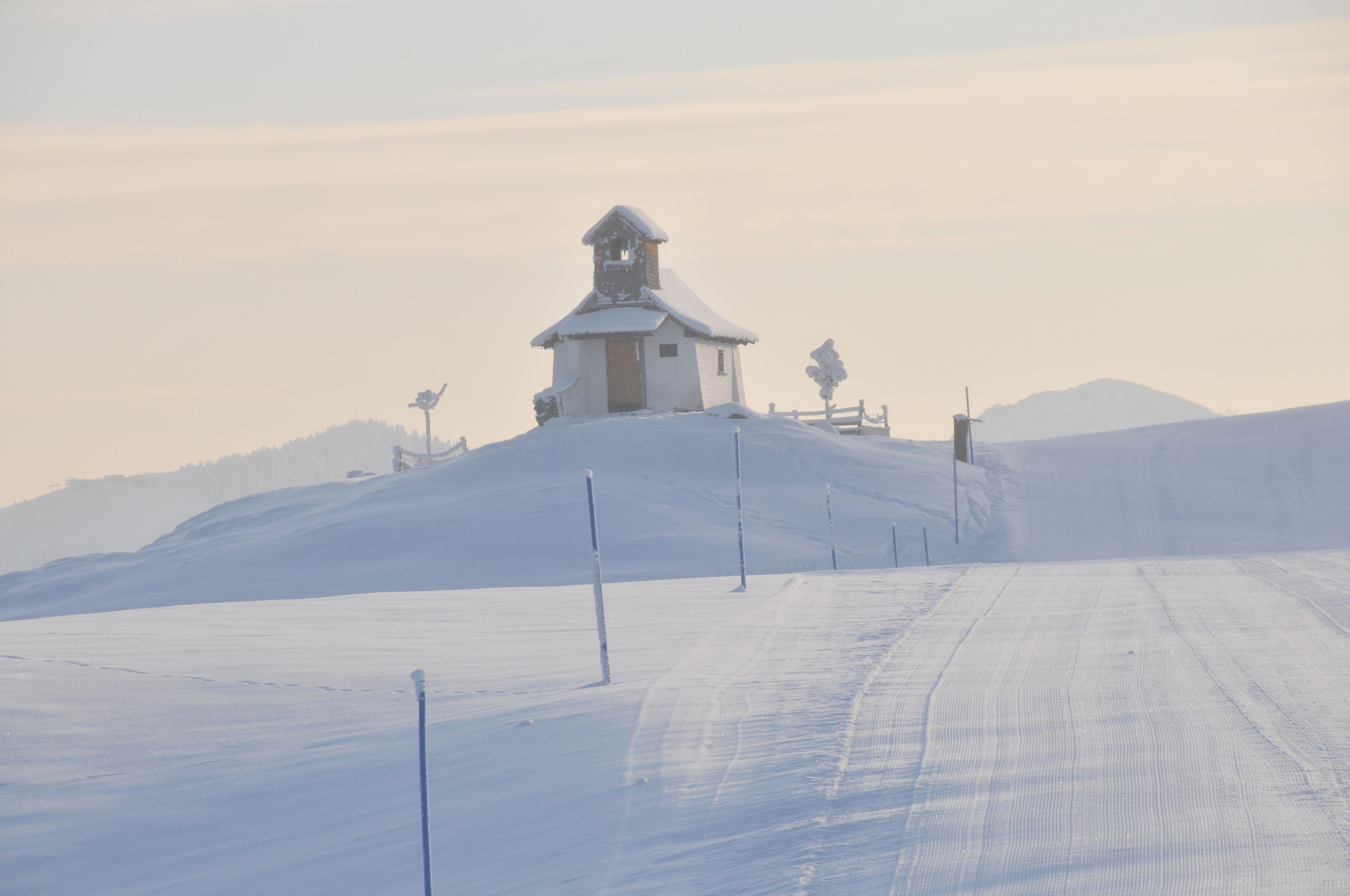 A snow-covered landscape with a small building on a hill, surrounded by snow and a clear sky.