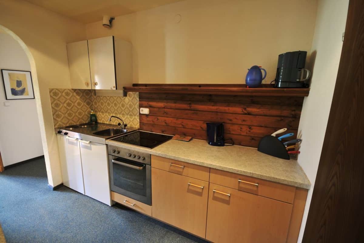 Una cocina compacta con armarios blancos, estufa, horno, lavamanos y cafetera sobre una repisa de madera.