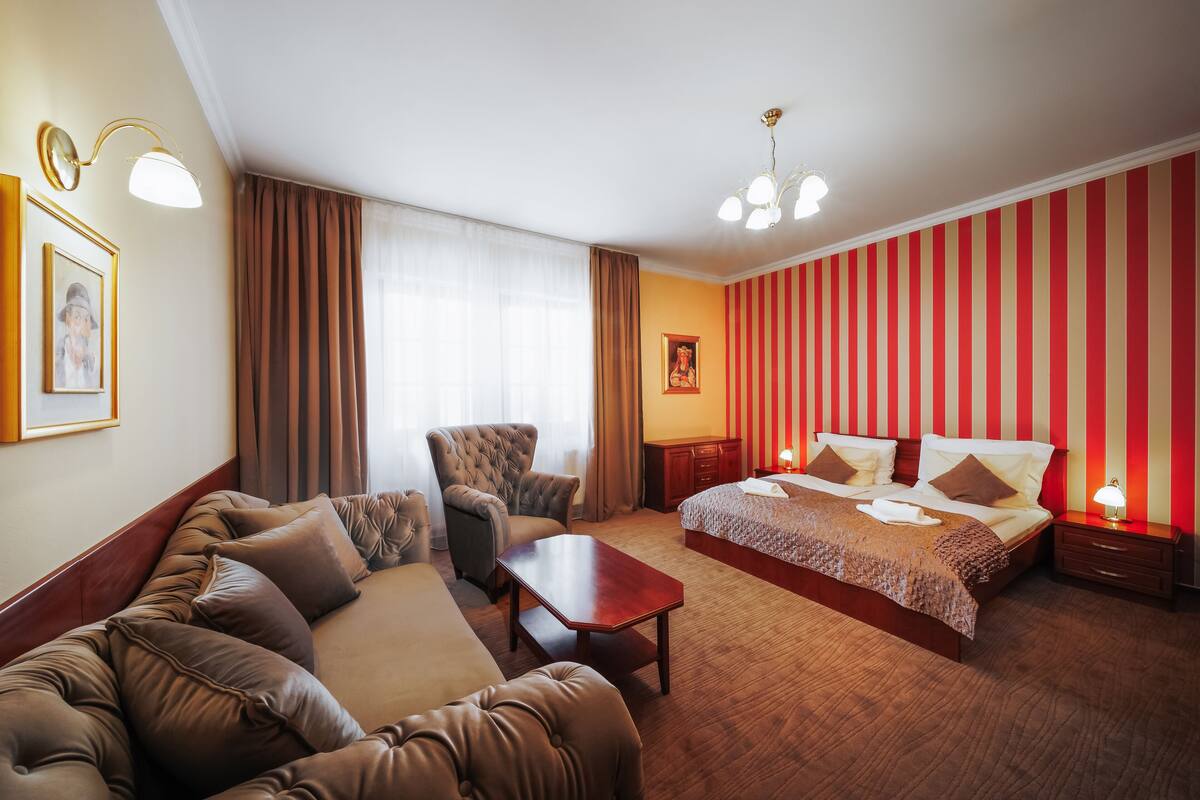 A hotel room with a large bed, a sofa, a chair, a small table, and a nightstand with a lamp.