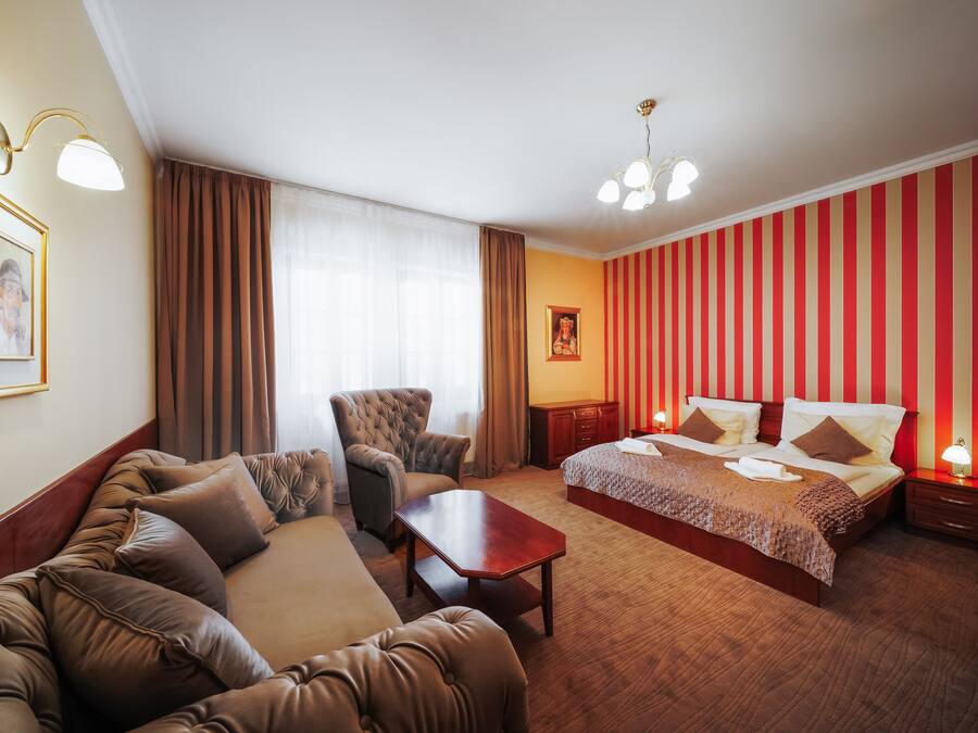 Una habitación de hotel con una cama grande, un sofá, una silla, una mesita pequeña y una mesita de noche con una lámpara.