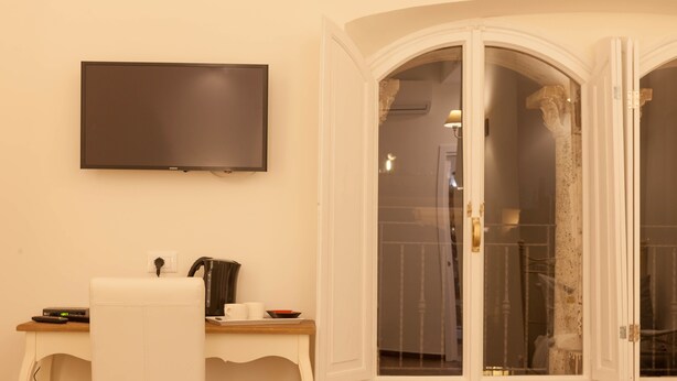 Una habitación con una televisión de pantalla plana montada en la pared, un escritorio blanco con una computadora, una silla y una ventana con vista a un baño.
