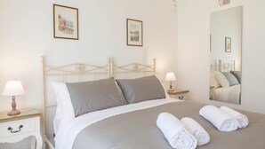 Una cama bien hecha con ropa de cama gris, dos lámparas de noche y un espejo que refleja la cama.