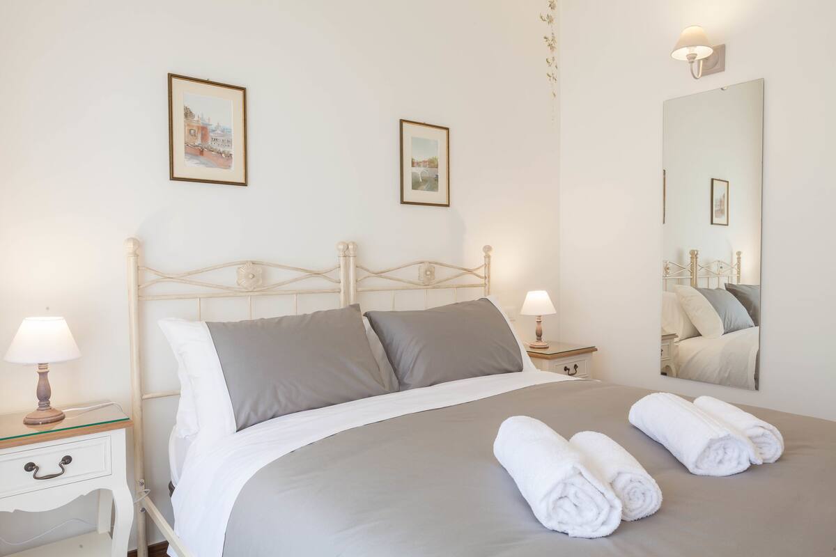 A neatly made bed with grey linens, two bedside lamps, and a mirror reflecting the bed.