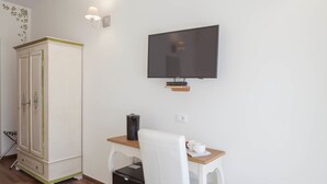 Una habitación pequeña con un escritorio de madera, una silla blanca, un televisor montado en la pared, un armario con paneles decorativos y un refrigerador negro compacto.