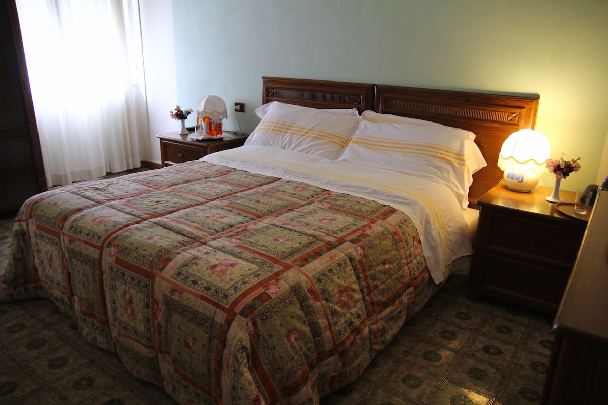 A neatly made bed with a floral-patterned bedspread, a wooden headboard, and a bedside table with a lamp and a vase of flowers.