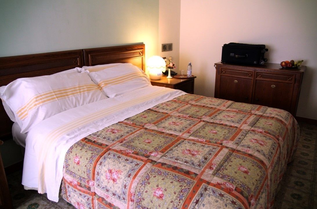 Una cama bien hecha con una colcha estampada de flores y almohadas blancas, una lámpara de noche, una mesita de noche de madera con una lámpara y un pequeño mueble de madera con una maleta y fruta encima.