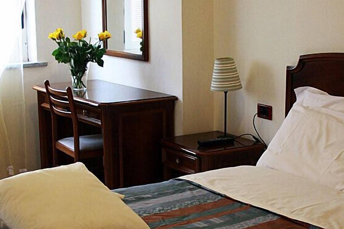 A hotel room with a bed, a bedside table with a lamp, a dresser, and a vase with flowers.