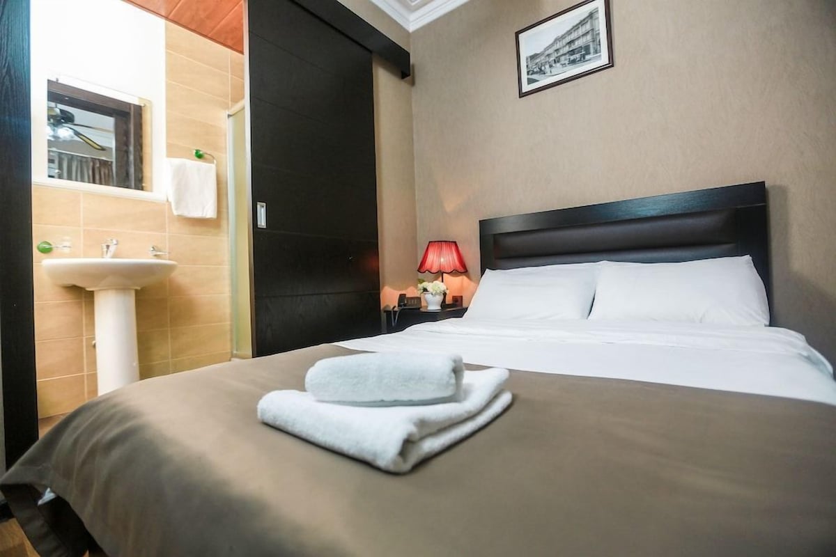 A hotel room with a bed, a nightstand with a lamp, a window, and a bathroom with a sink and mirror.