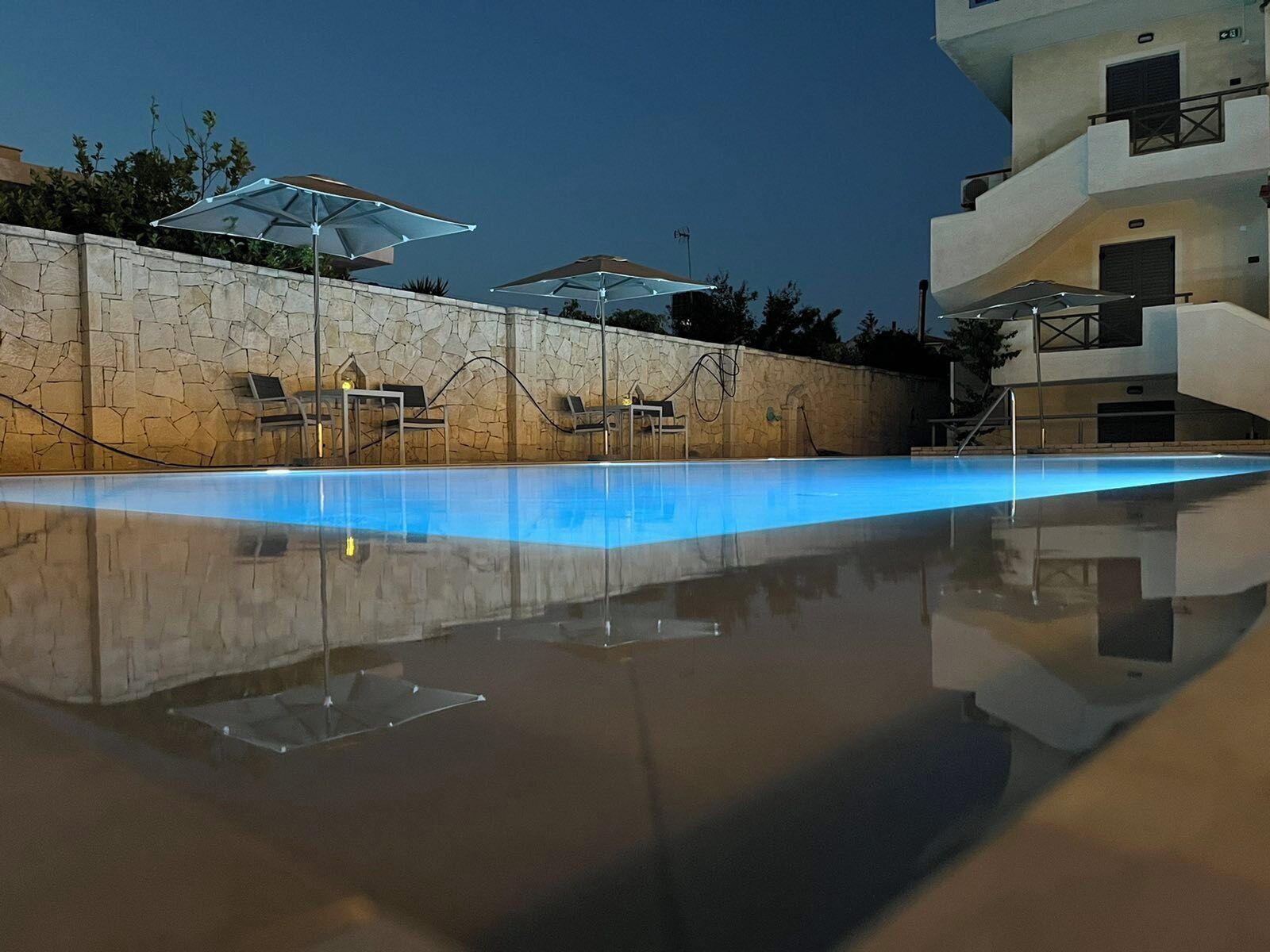 Une piscine entourée de chaises longues et de parasols, le tout bordé de murs en pierre et adossé à un bâtiment moderne.
