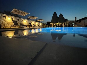 A pool with lounge chairs and umbrellas, stone walls, and a building with a unique architectural design. - Nancy - Chara Apartments (Heraklion)