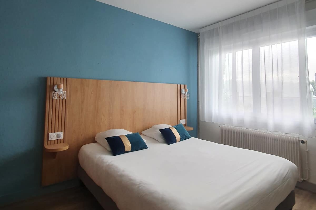 A bedroom with a wooden headboard, a large bed with white bedding, and a window with sheer curtains.