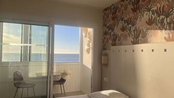 Een slaapkamer met een bed, een stoel, een klein tafeltje en uitzicht op zee door een schuifdeur.