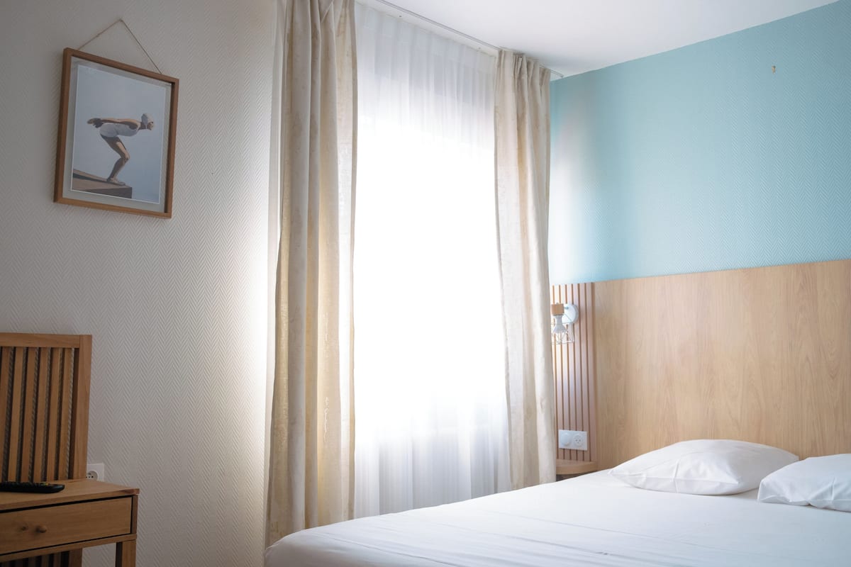 A hotel room with a bed, a wooden nightstand, a framed picture on the wall, and a window with curtains.