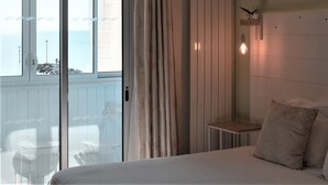 A bedroom with a bed, a small table, a nightstand, a wall-mounted light, and a view of the sea through a sliding door. - Hôtel Le Rivage (Chatelaillon-Plage)