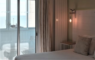 Quarto com cama, mesinha, mesa de cabeceira, luminária na parede e vista para o mar através de uma porta deslizante.