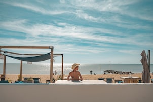 Vista da praia com uma rede, espreguiçadeiras e uma mesa sob área sombreada.