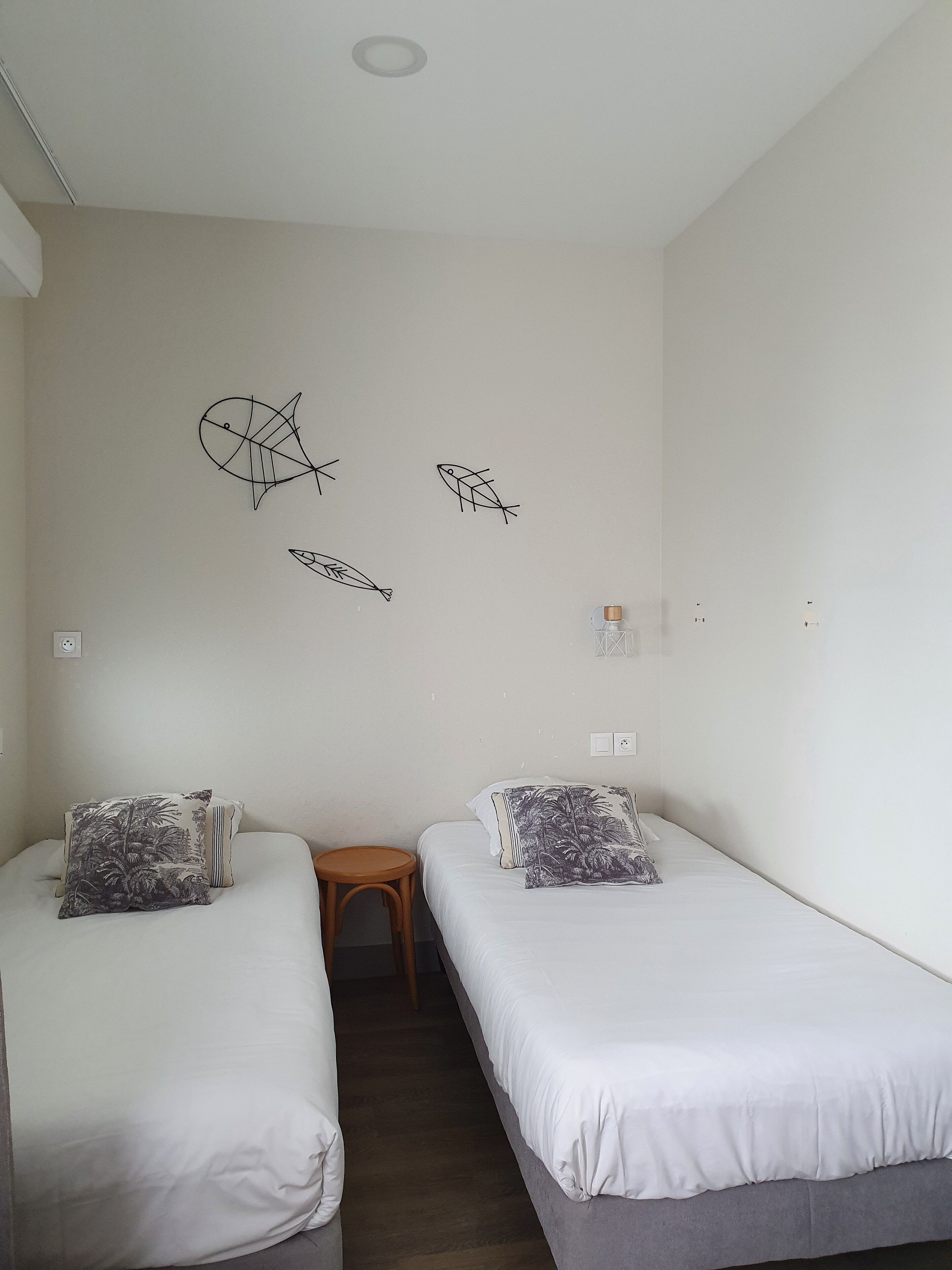 Ein Zimmer mit zwei Betten, jeweils mit Kissen und einem kleinen Holzschemel. An den Wänden befinden sich Fischdarstellungen und ein Lichtschalter.