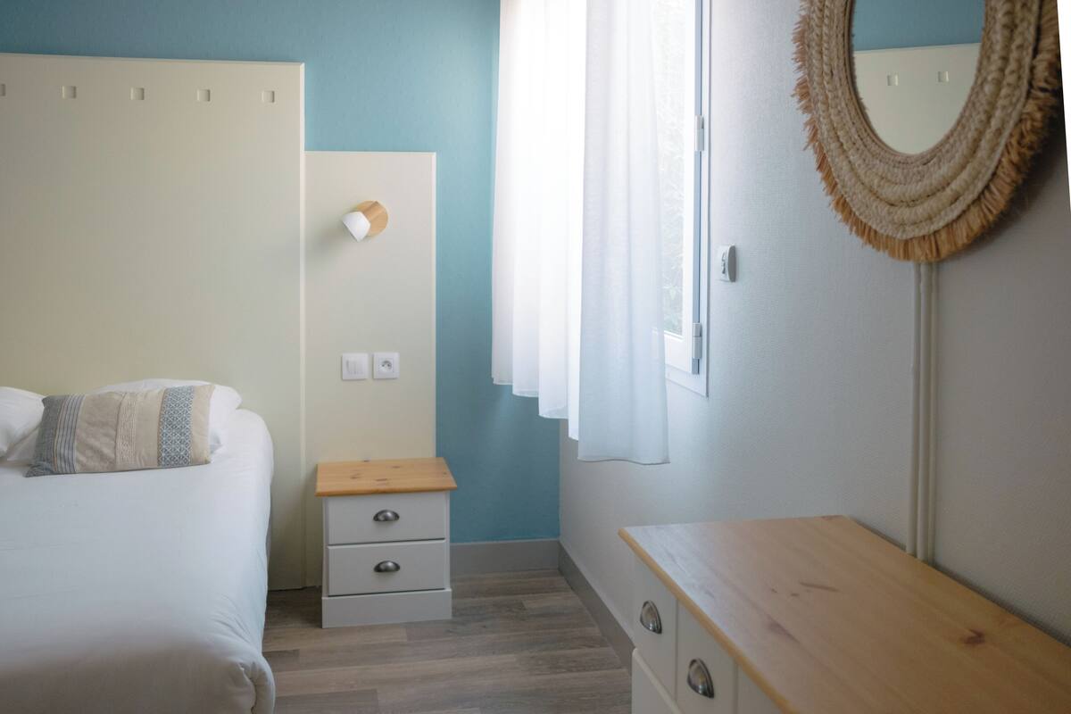 A bedroom with a bed, a bedside table, a dresser, a mirror, and a window with curtains.