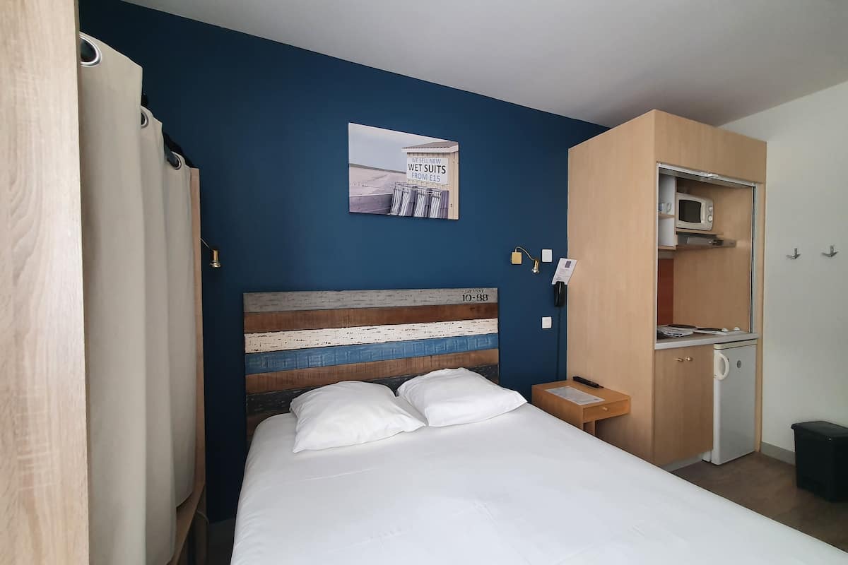 A bedroom with a bed, a wooden headboard, a small bedside table, a nightstand, a wall-mounted telephone, and a kitchenette with a microwave and a small refrigerator.