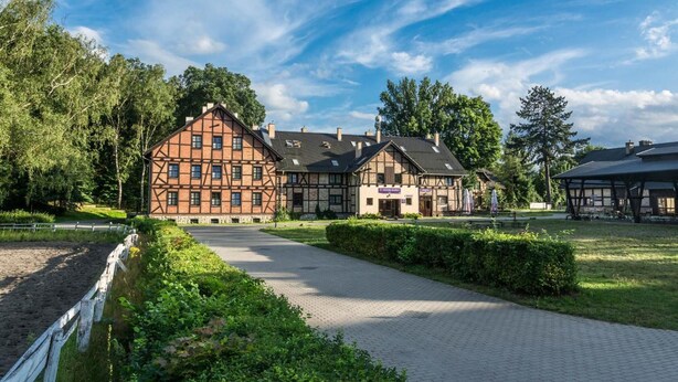 A traditional half-timbered building with a paved pathway leading to it, surrounded by greenery and trees. - Folwark Książęcy (Osiecznica)
