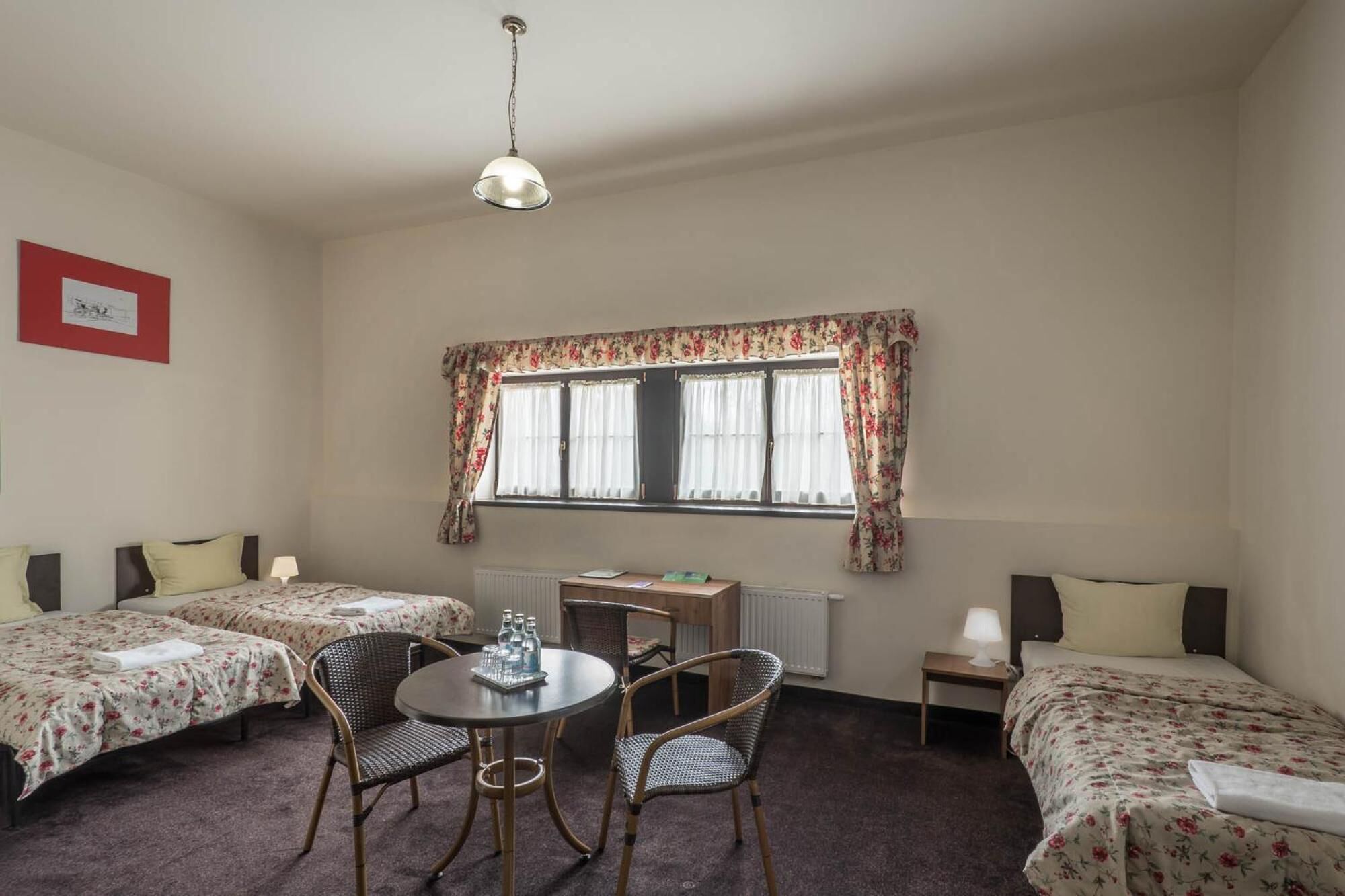 A room with two single beds, a round table with chairs, a small table with a lamp, and a window with floral curtains.