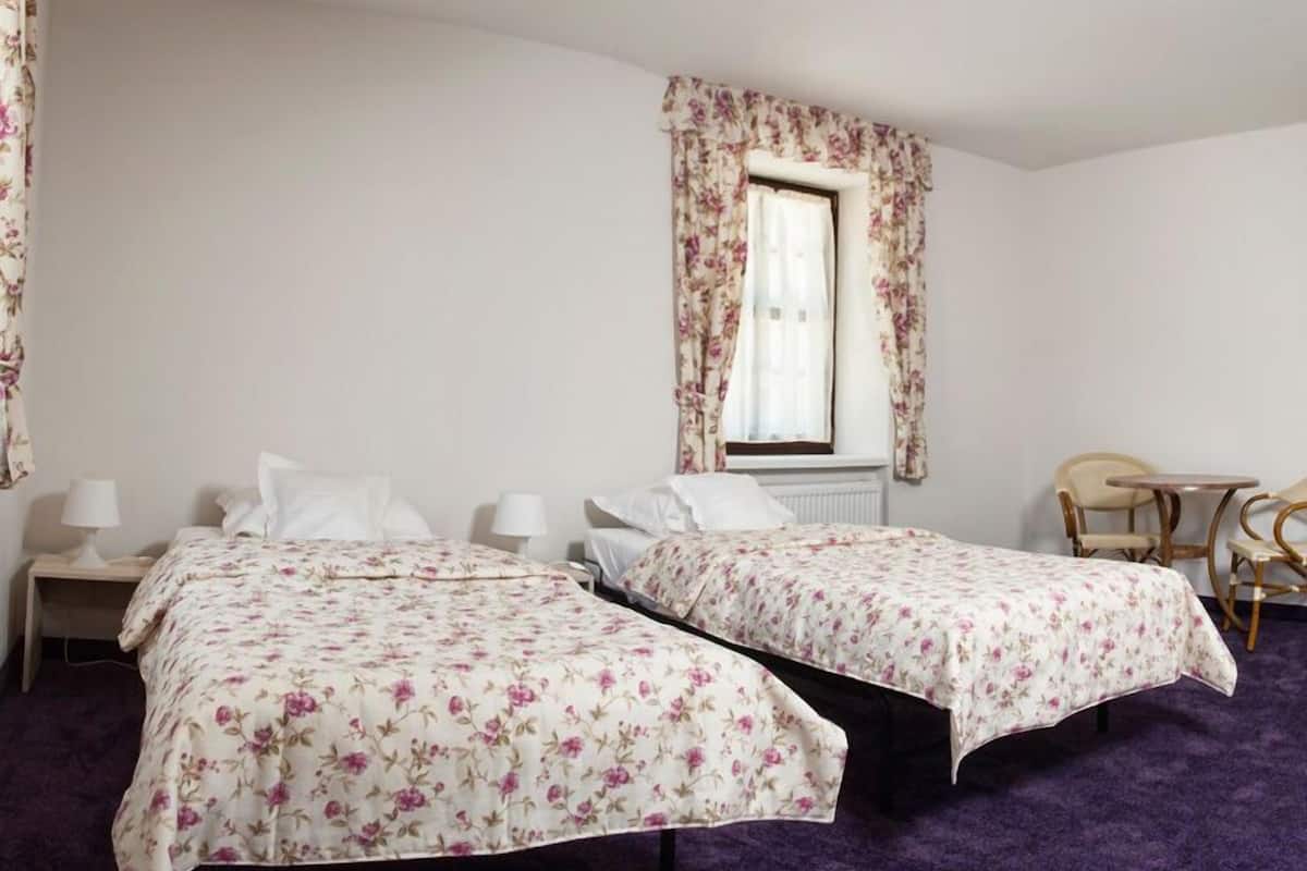 A hotel room with two single beds, a small table, two chairs, a window with floral curtains, and a radiator.