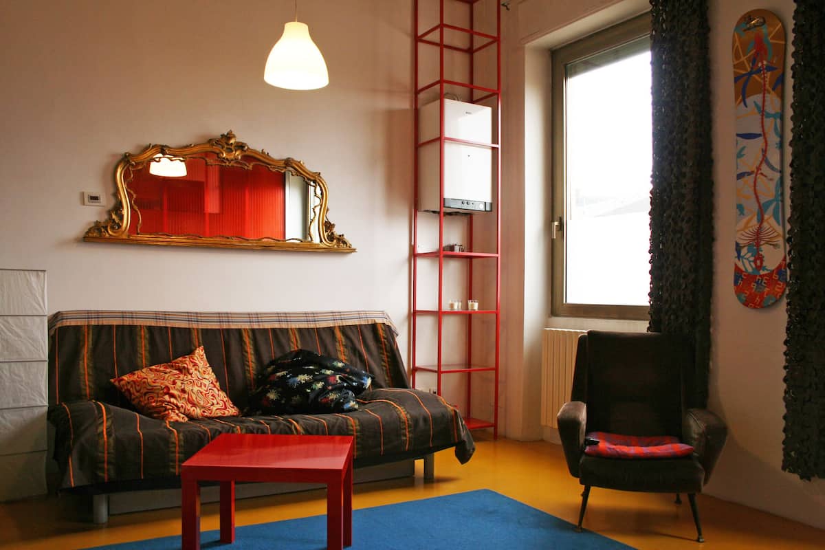 A living room with a sofa, a red table, a chair, a mirror, a radiator, and a window with curtains.