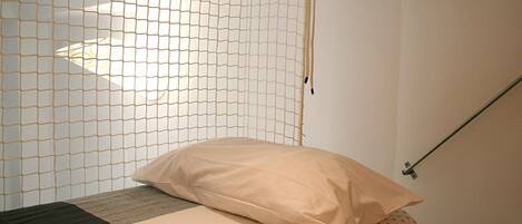 Una cama con colchón color beige y una sola almohada, una ventana con mosquitero y una barra de metal.