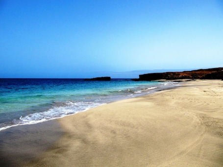 A beach with clear blue water, sandy shore, and rocky cliffs in the background.