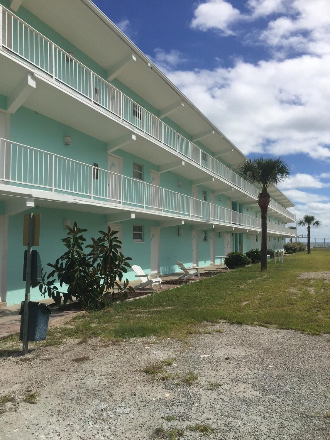 A multi-story hotel with a light blue exterior, white railings, and a covered outdoor seating area with lawn chairs.