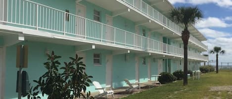 A multi-story hotel with a light blue exterior, white railings, and a covered outdoor seating area with lawn chairs.
