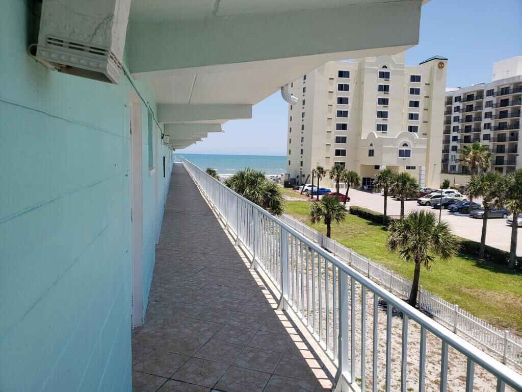 Een balkon met uitzicht op het strand en de oceaan, een parkeerplaats met auto's, en een gebouw met meerdere verdiepingen.
