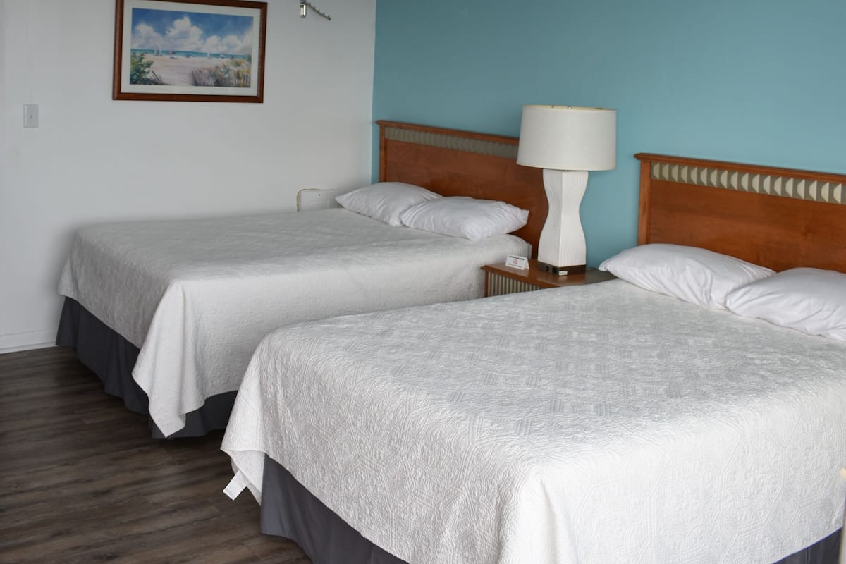A hotel room with two beds, a nightstand with a lamp, a painting on the wall, and a wooden floor.