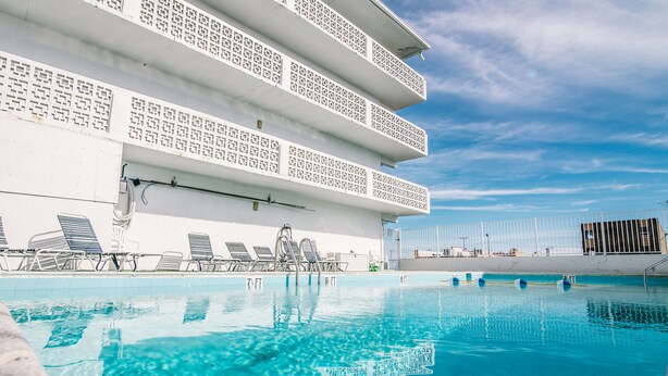 A swimming pool with clear blue water, lounge chairs, and a white building with patterned balconies.