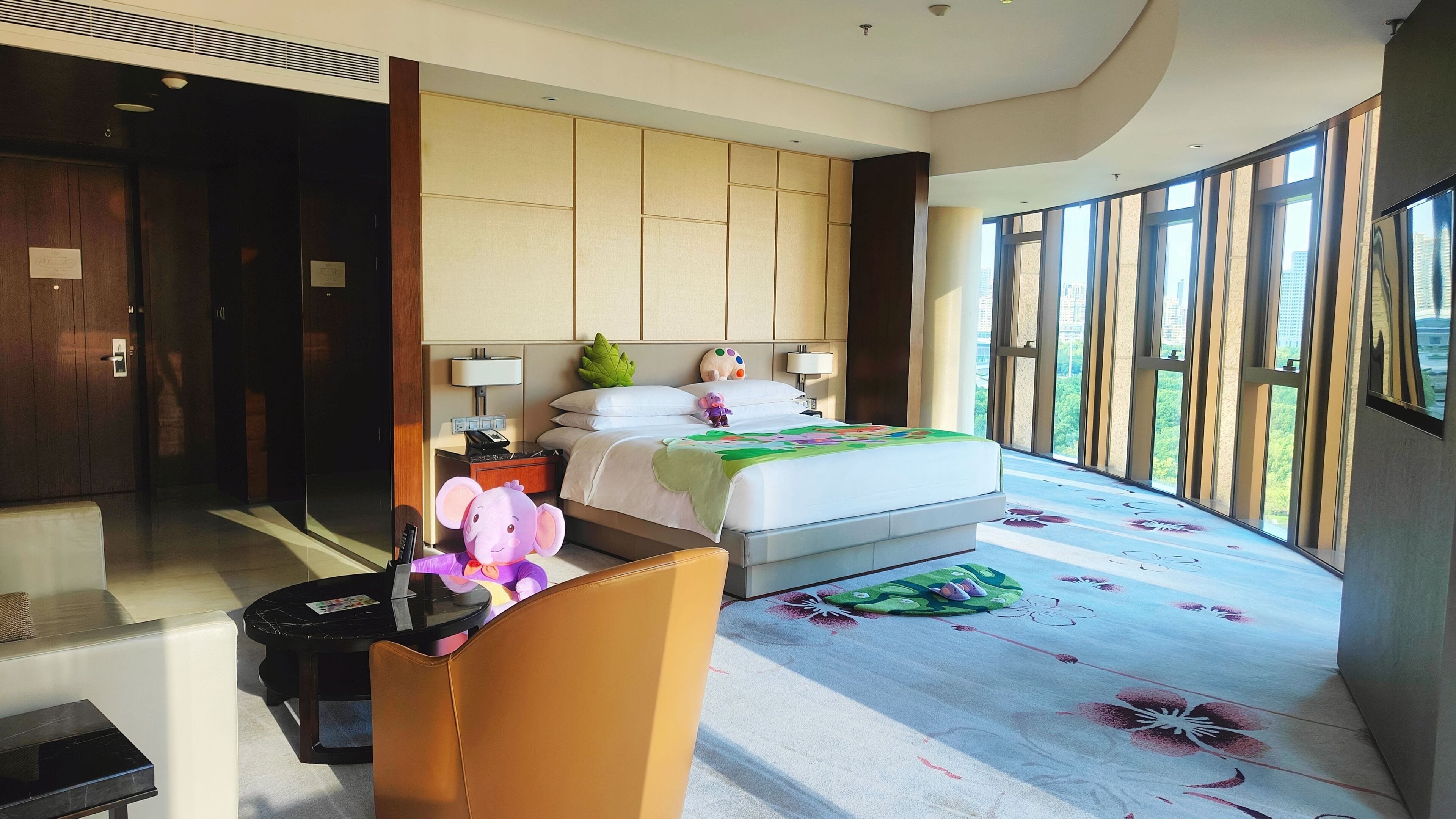 A hotel room with a bed, a chair, a desk, and a large window offering a city view.
