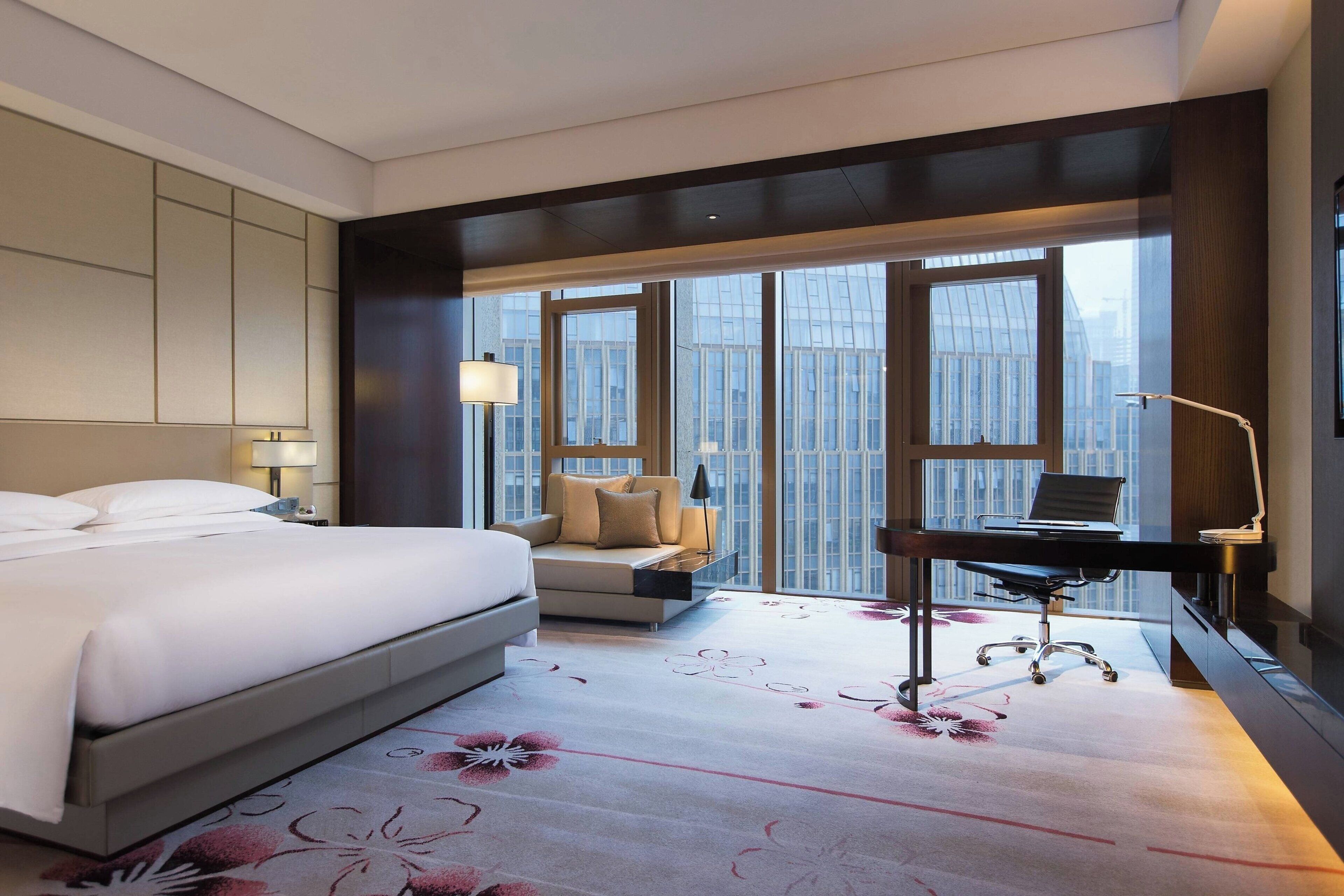 A hotel room with a bed, a desk, a sofa, and large windows offering a city view.