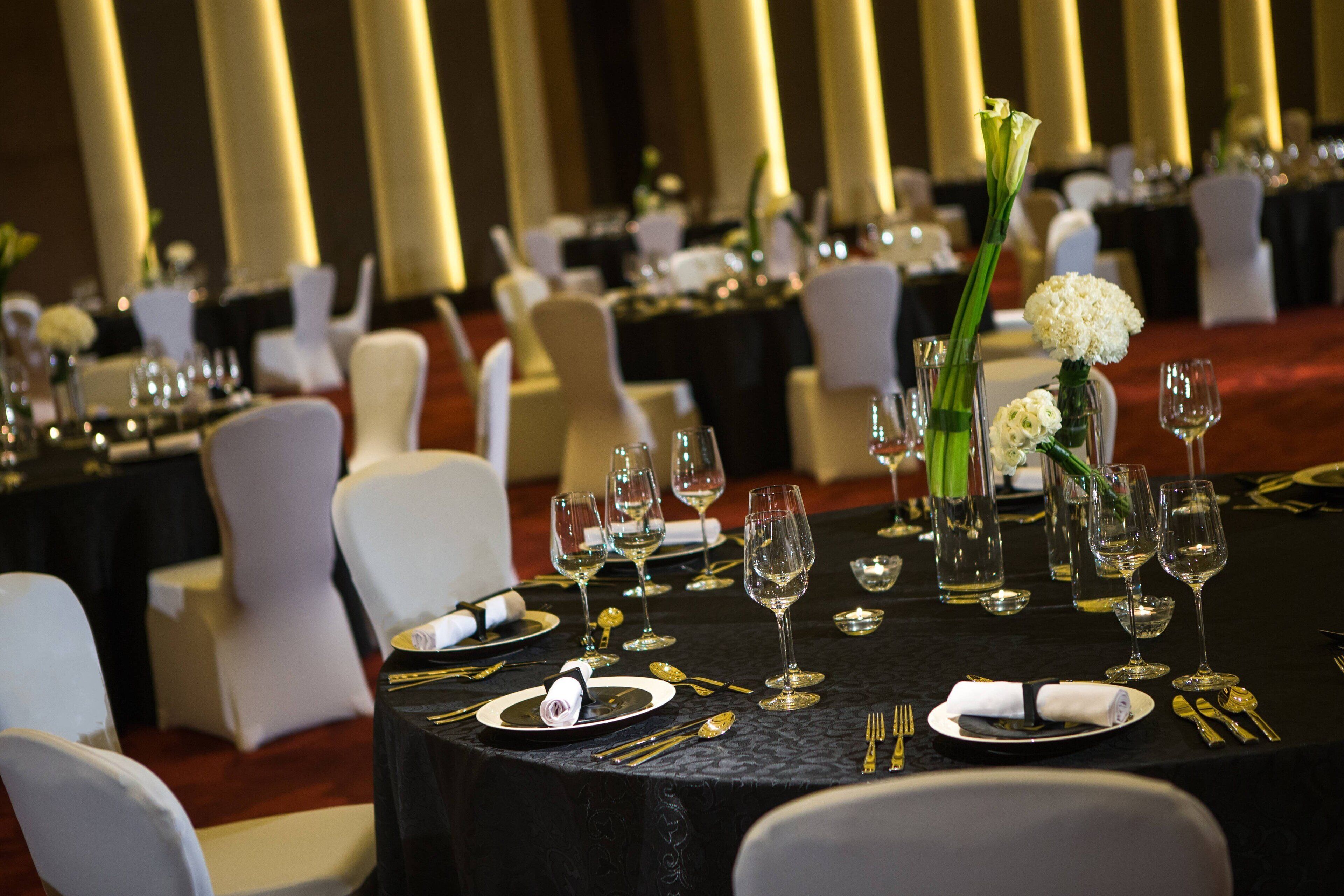A banquet hall with round tables set for dining, each with white chairs, black tablecloths, and gold cutlery.