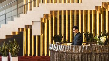 A reception desk with a marble top, a man standing behind it, and a staircase with a modern design in the background.
