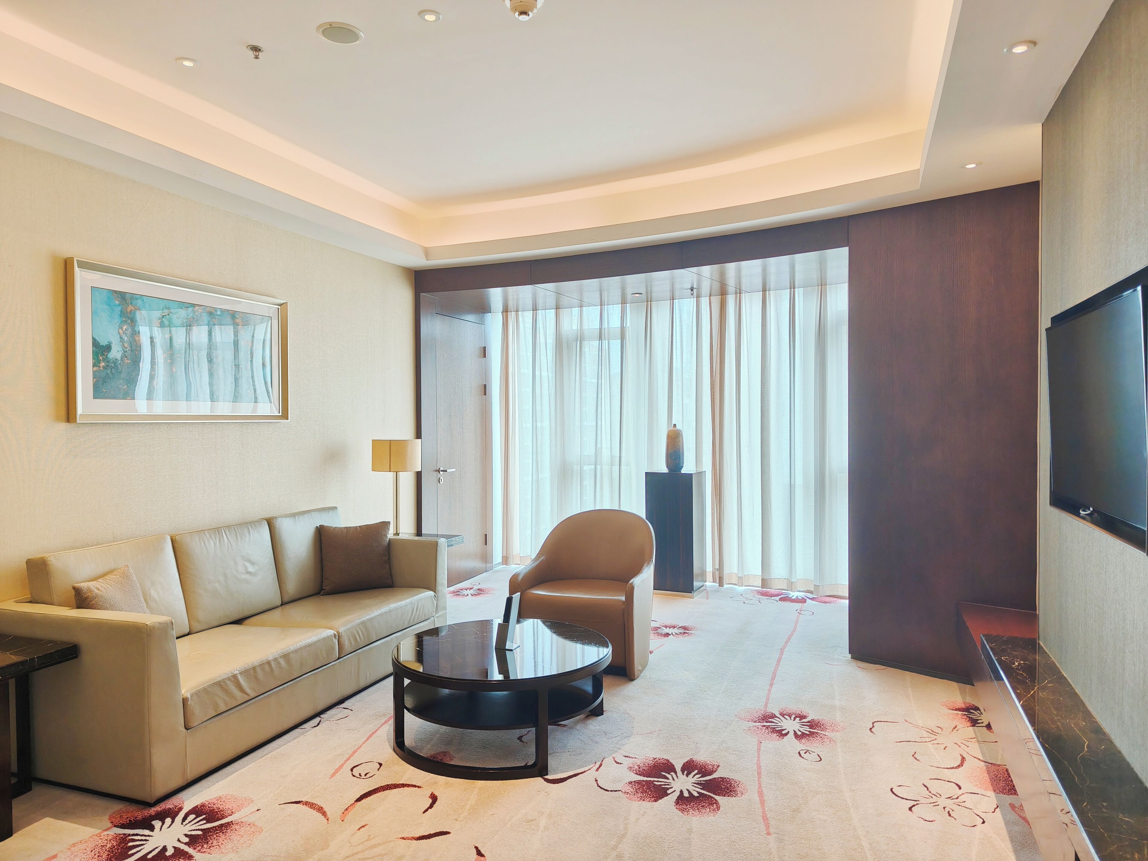 A hotel room with a sofa, a round coffee table, a TV, and a large window with curtains.