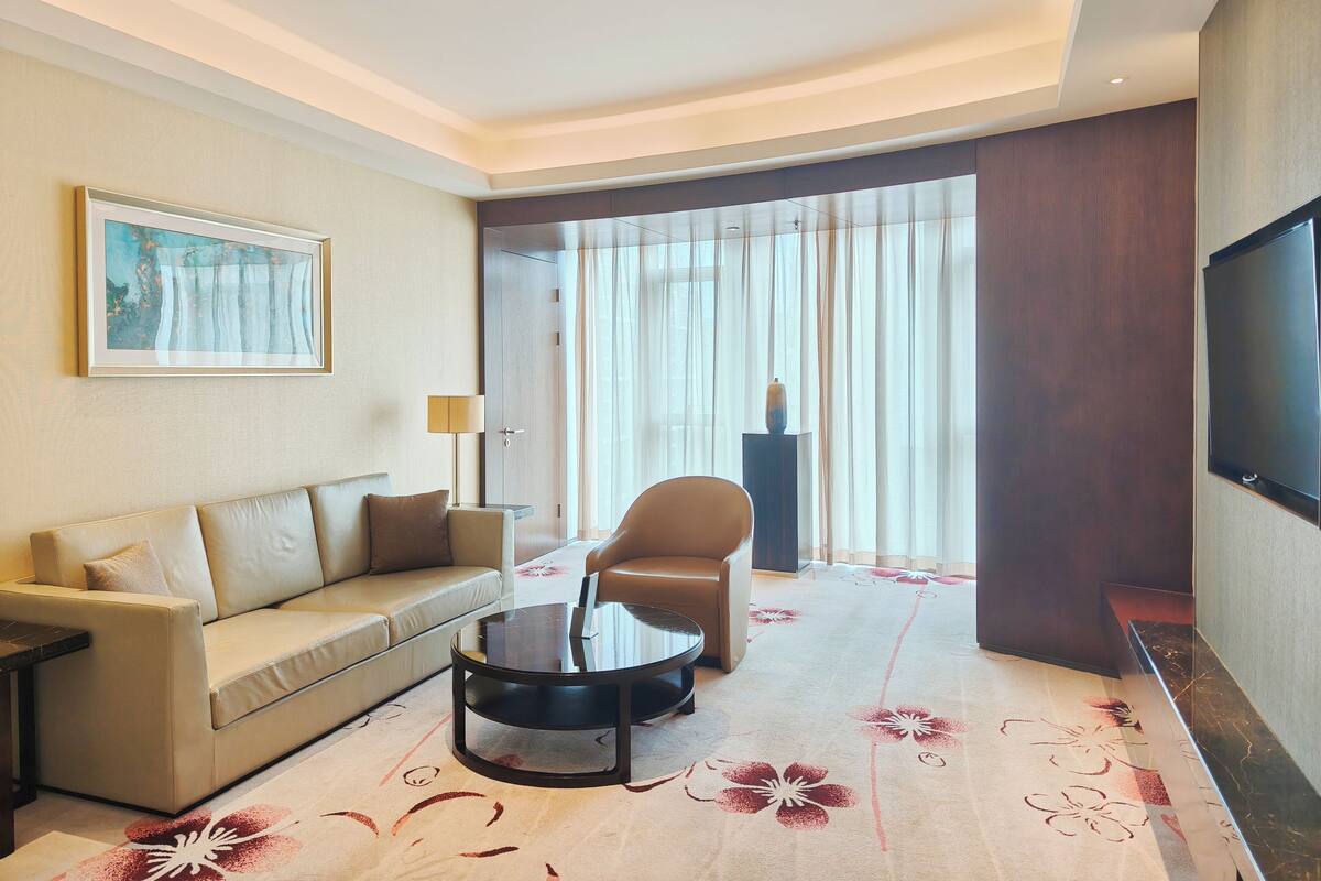 A hotel room with a sofa, a round coffee table, a TV, and a large window with curtains.