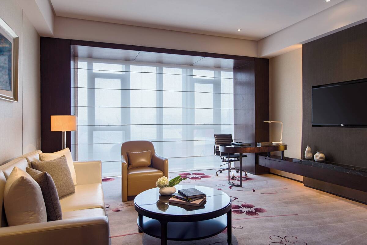 A modern hotel room with a large window, a sofa, a round coffee table, a desk, and a flat-screen TV.