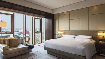 A hotel room with a large bed, a sofa, a nightstand, and a view of the cityscape through large windows.