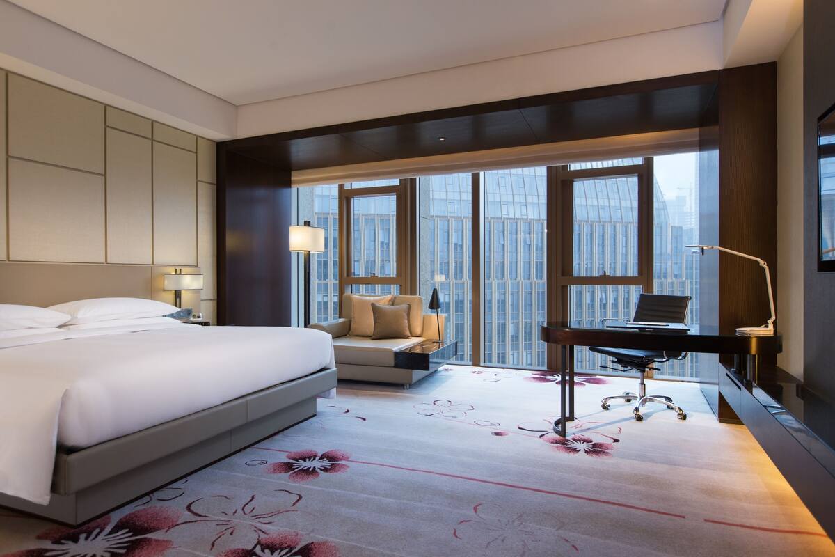 A hotel room with a large bed, a desk, a chair, and a view of the city through large windows.