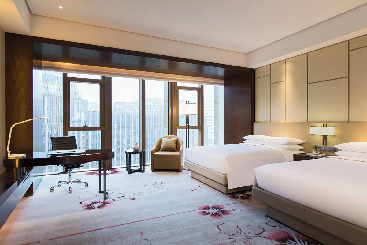 A hotel room with a desk, two beds, a chair, and a large window with a city view.